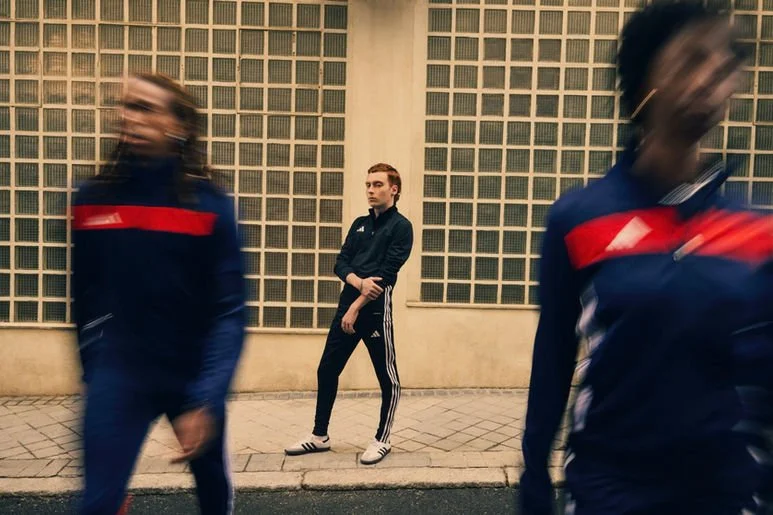 A young man stands against a yellow wall with glass blocks, while two blurred individuals walk past him in the foreground.