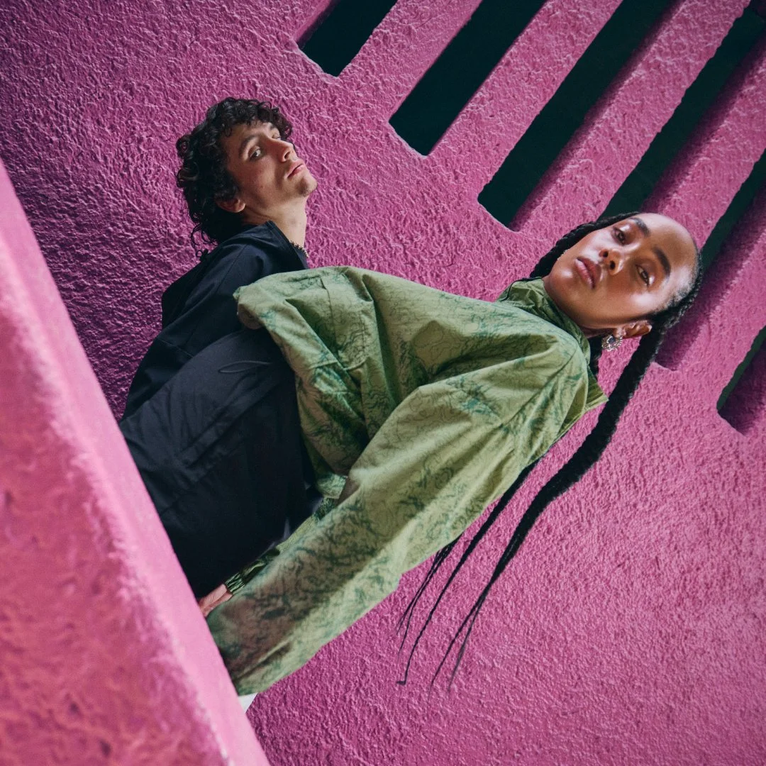 Two people standing against a textured pink wall with black slats.