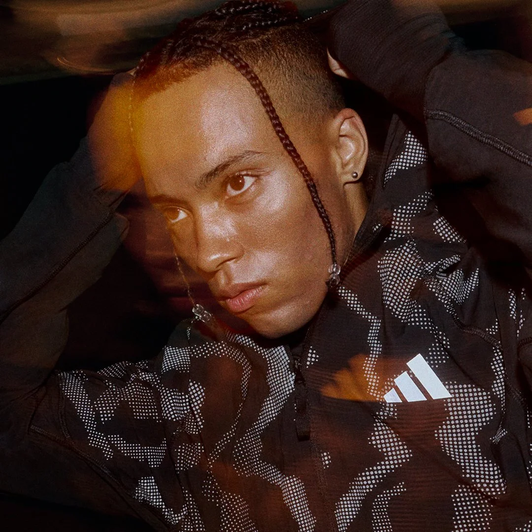 Close-up of a young person with braided hair, lying down with hands behind head, wearing a black and white patterned Adidas jacket, in a dimly lit setting.
