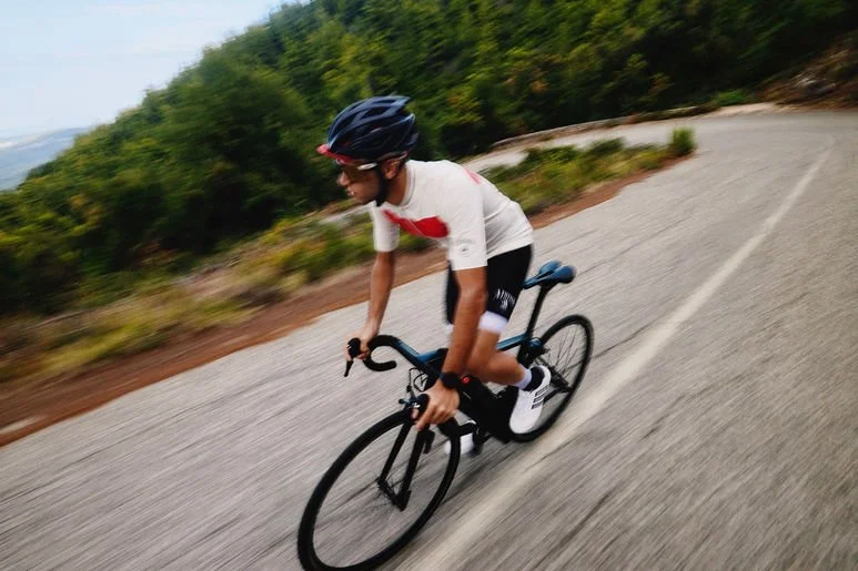 Person riding a black road bike on a winding mountain road, wearing a helmet, white cycling jersey with red accents, and black shorts, with greenery and trees in the background.