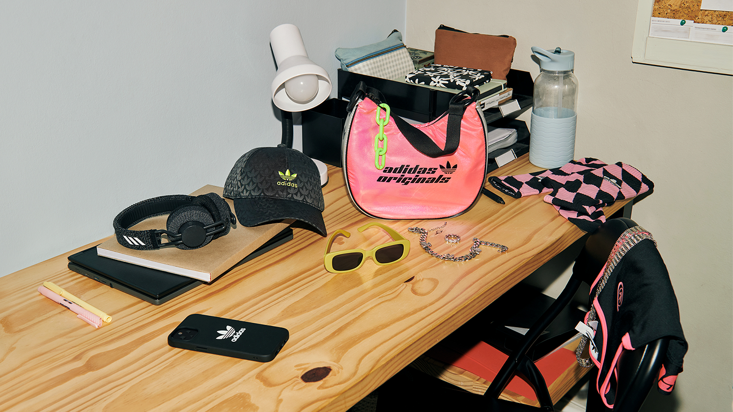 Cluttered wooden desk with various items including a black Adidas hat, black headphones, a black phone with Adidas logo, yellow sunglasses, pink Adidas bag, skullcandy case, jewelry, notebooks, a pink pen, and a chair with a black hoodie that has pin