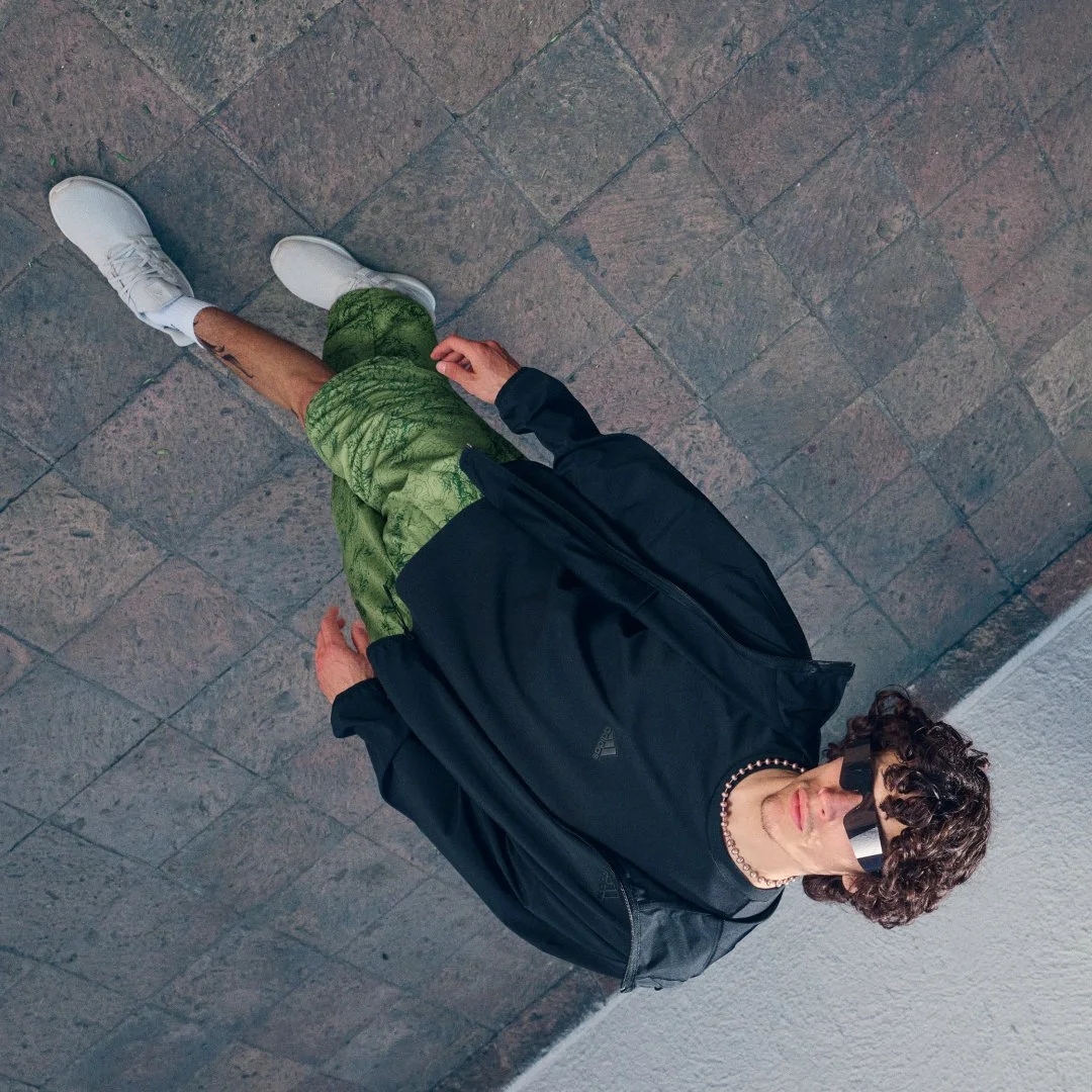 A person with curly hair wearing sunglasses, a black jacket, a black choker, and green shorts, sitting on the ground and leaning against a wall.