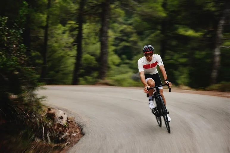 A person riding a bicycle on a curved forest road, wearing a helmet and athletic clothing.