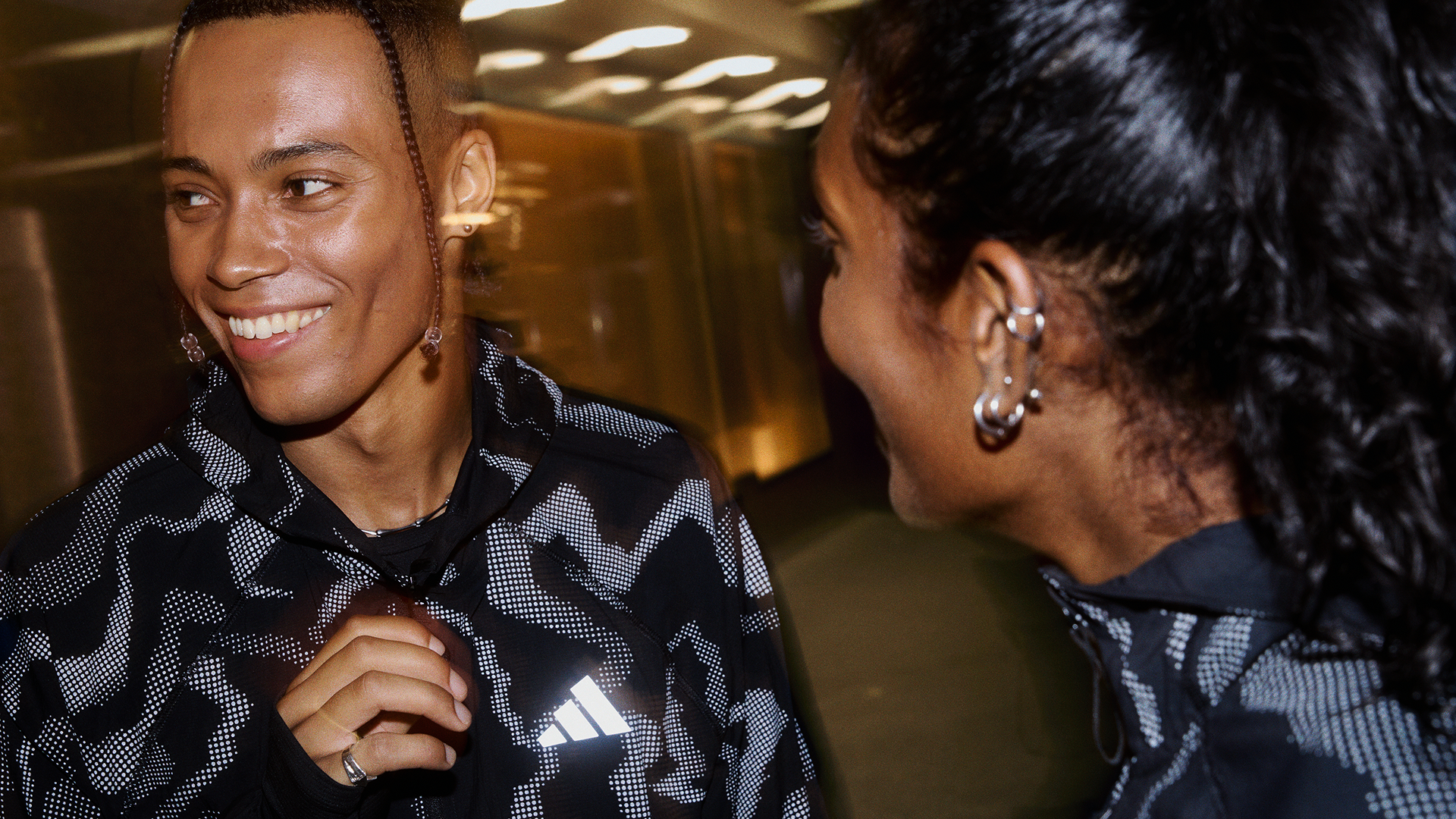Two women sharing a friendly conversation indoors, one smiling and the other with her head turned towards the first, both wearing athletic jackets with a black and white pattern.
