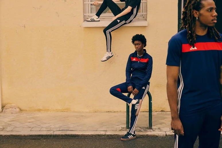 Three people in matching Adidas sportswear, two in blue and red track suits, with one sitting on a green bike rack and the other standing on a sidewalk, and the third seated on a window ledge on top of a yellow wall.