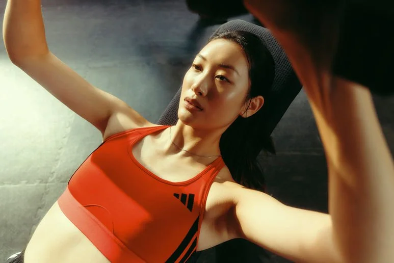 A young woman in an orange sports bra lying on a gym mat during workout, with a focused expression, resting on a workout bench.