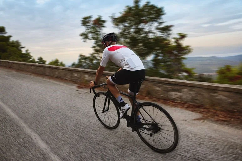 A person riding a black road bike on a winding paved road with a safety helmet, wearing a white and red cycling jersey, black shorts, and dark shoes, with blurred trees and landscape in the background.