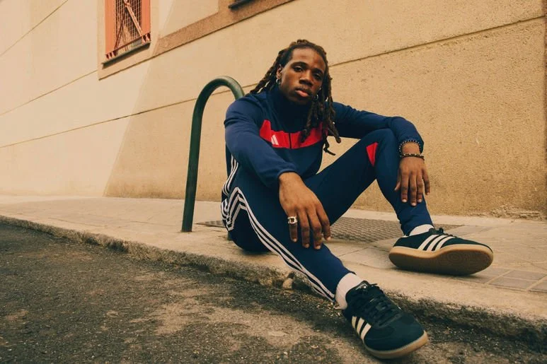 A young man with dreadlocks sitting on a sidewalk next to a beige building, wearing a navy blue athletic tracksuit with red and white accents and black and white sneakers.