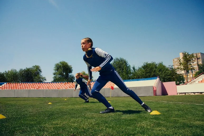 Two female athletes in athletic gear performing sprint drills on a grass field marked with small yellow cones under a clear blue sky.