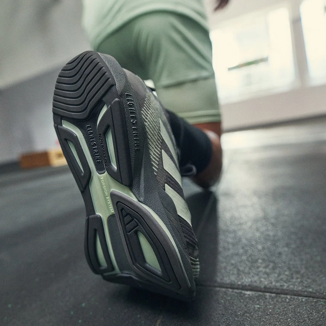 Close-up of athletic shoe sole on gym floor, person in background wearing green shirt and dark shorts