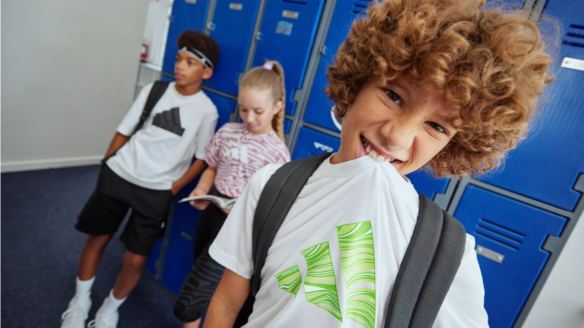 A smiling boy with curly hair in the foreground holding a backpack, with two other children, a boy and a girl, standing near blue lockers in the background, in a school hallway.