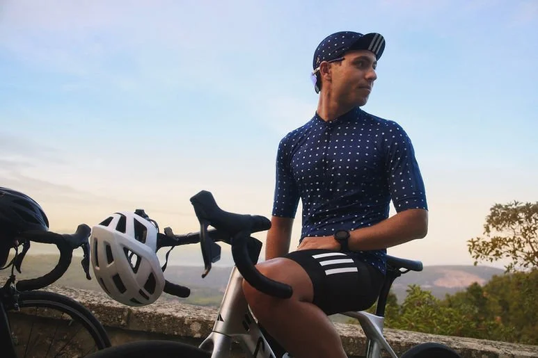 A man in cycling gear, including a polka-dotted helmet and jersey, sitting on a bicycle with a scenic background of trees and hills during sunset or sunrise.