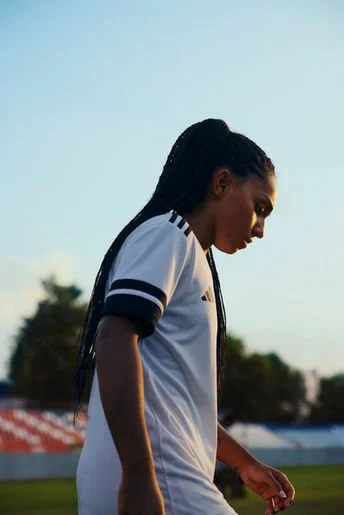 A young woman with braided hair in a sports jersey on a sports field with stadium seating and trees in the background.