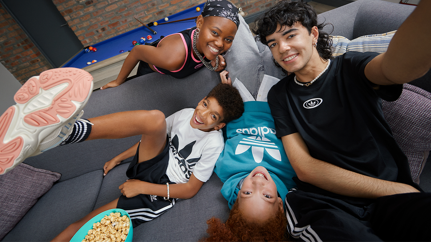 Group of four kids smiling and taking a selfie on a gray couch in a game room, with a pool table in the background, popcorn in a blue bowl, wearing casual sporty clothes.