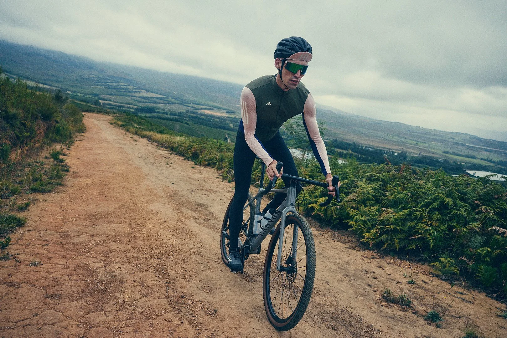 A man wearing a helmet, sunglasses, and cycling gear riding a gravel bike on a dirt trail surrounded by greenery with a scenic landscape and cloudy sky in the background.