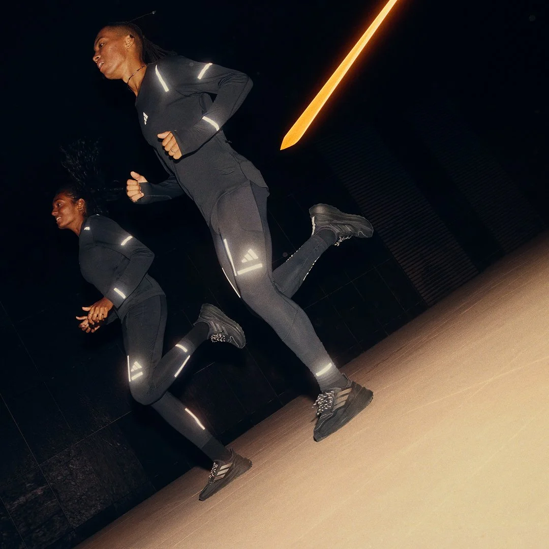Two women running indoors in black athletic clothes with reflective detailing, on a dark background with a yellow light streak.