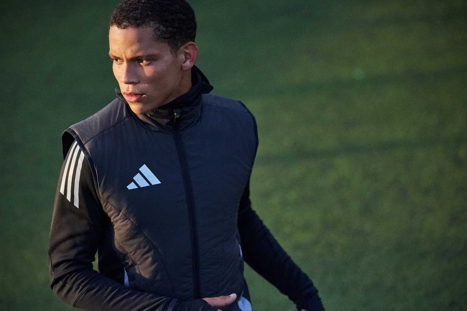 A man in a black athletic jacket with white stripes on the sleeves standing on a grassy field.