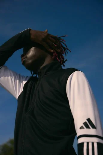 Person wearing a black and white athletic jacket with Adidas logo, holding hand above face against a blue sky.