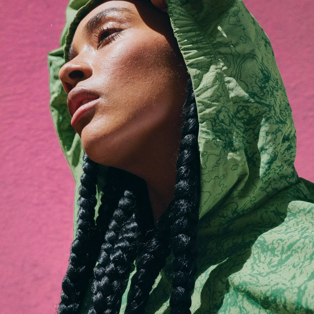 A woman with dark braided hair looking upward, wearing a green hooded jacket with a pattern, against a pink background.