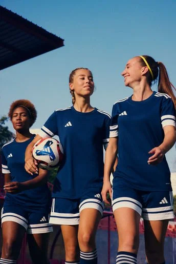 Three female soccer players in blue uniforms on the field, one holding a soccer ball, smiling and talking.