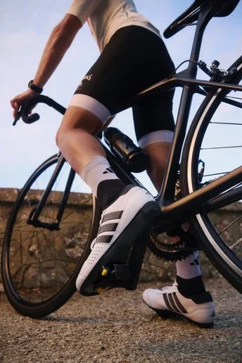 Close-up of a cyclist's legs and bike, showing black and white cycling shoes, white socks with black stripes, and a black road bike with a water bottle.