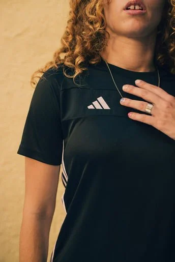 Close-up of a woman with curly hair wearing a black Adidas sports shirt, with a hand on her chest and a ring on her finger.