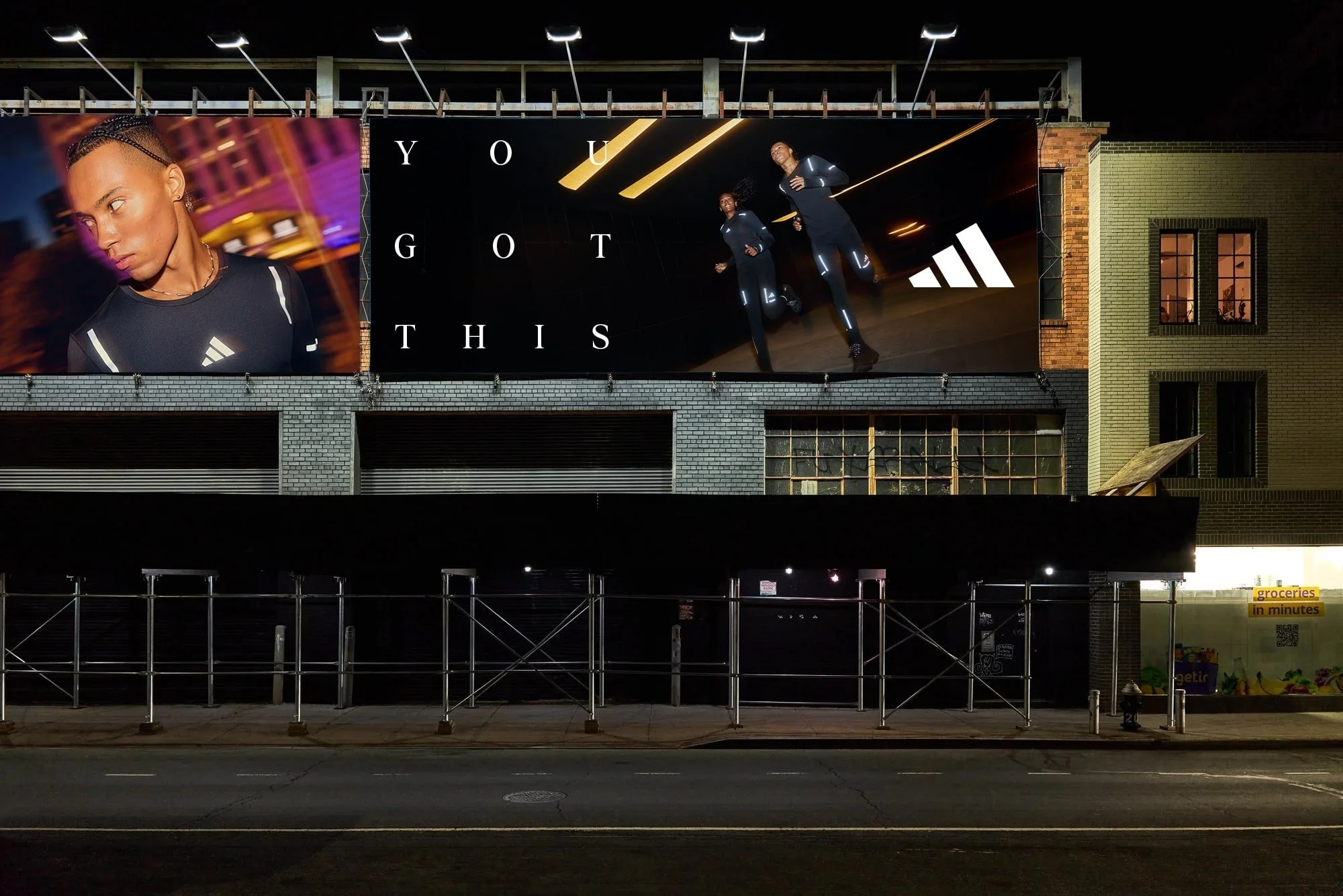 A large outdoor advertisement featuring two women running in a futuristic tunnel, wearing athletic clothing with bright lines and the phrase "YOU GOT THIS" displayed across the top. The building has a brick facade and a small window on the right, with a grocery store sign on the lower right corner.