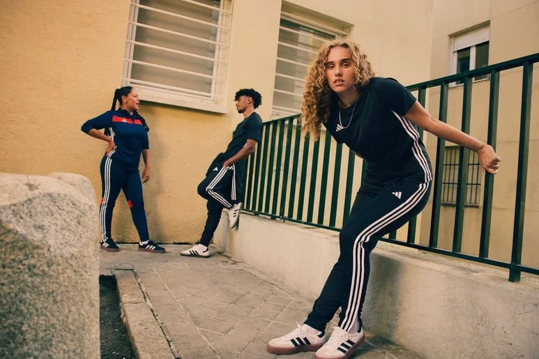 Three teenagers in sporty outfits with Adidas stripes, standing and leaning against a black railing outside a building with a yellow wall and white windows.
