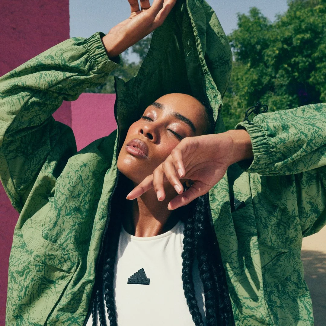 A woman wearing a green patterned jacket and a white Adidas shirt with braided hair, posing outdoors with her eyes closed and one hand near her face.