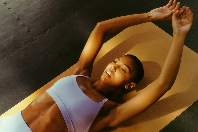 A woman lying on a yoga mat performing an abdominal stretch with arms extended overhead in a workout space.
