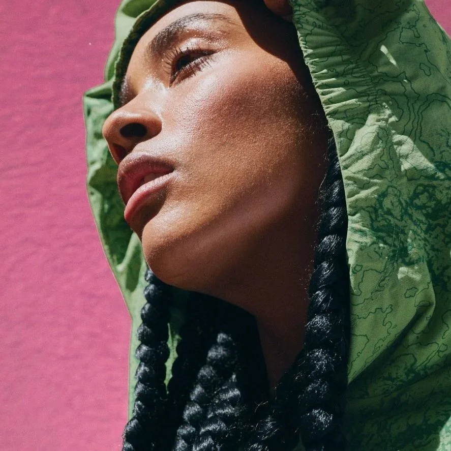 Close-up of a woman wearing a green hooded jacket with topographical patterns, against a pink background.