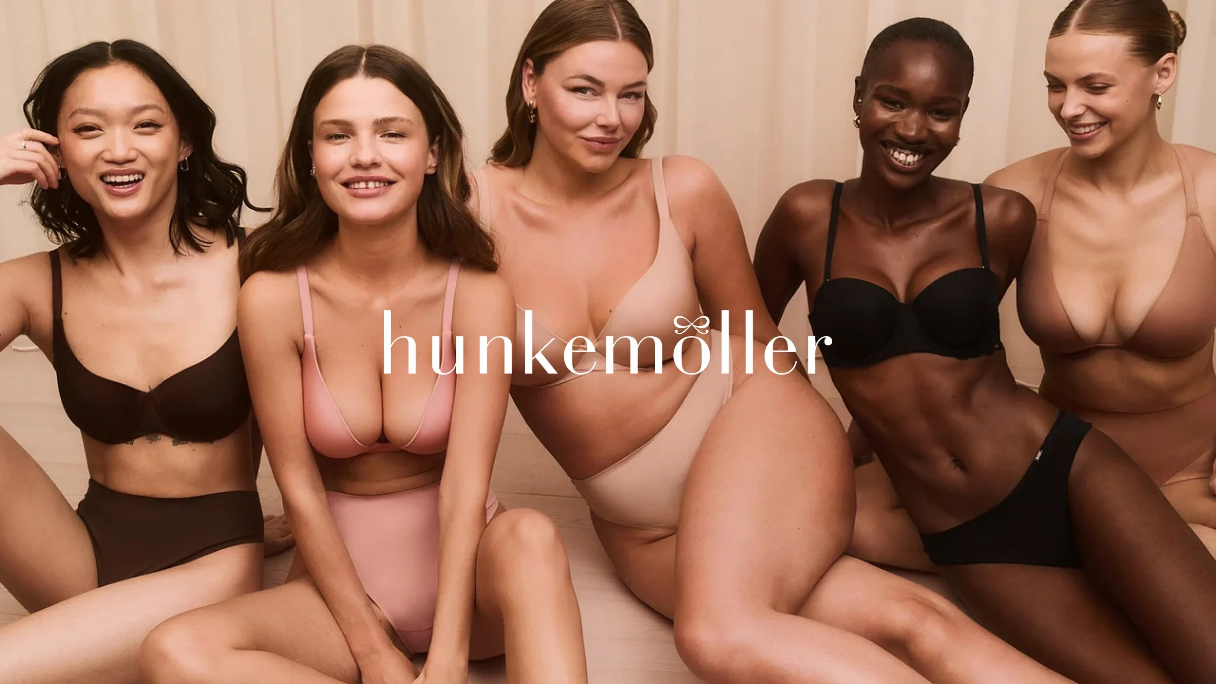 Group of five diverse women sitting together, wearing bras and underwear, smiling and looking at the camera.