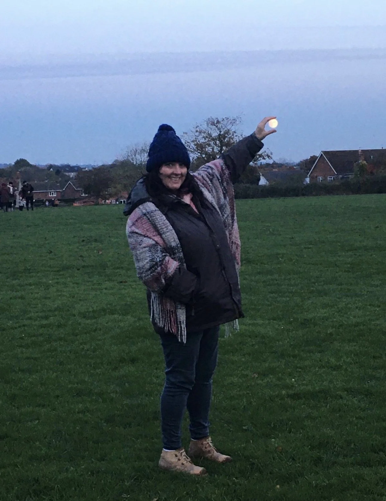 A woman standing on a grassy field at dusk, smiling and holding a small glowing orb in the air.