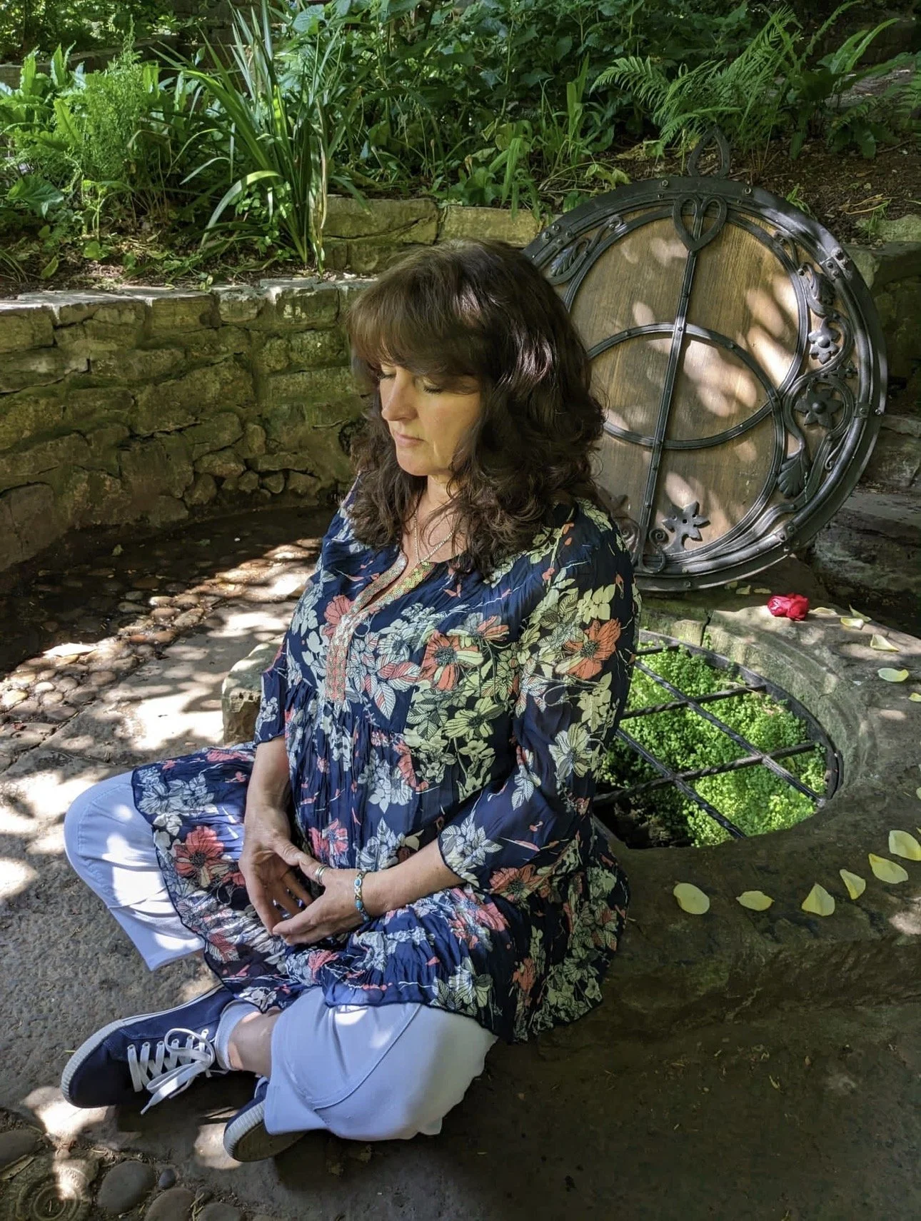 A woman with brown curly hair and a floral dress sitting in a garden, with a large decorative clock behind her and green plants around.