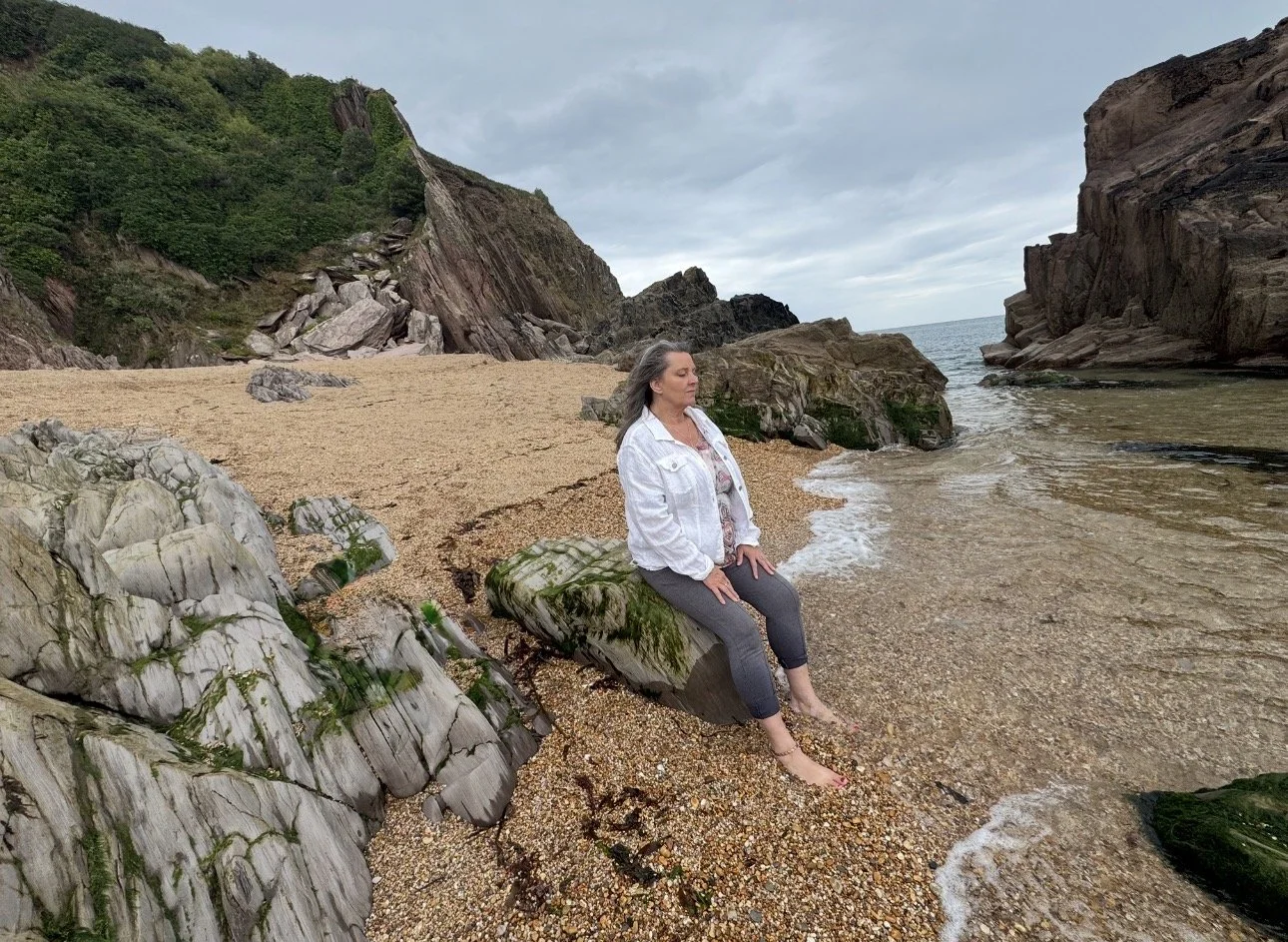 A woman sitting on a moss-covered rock at a beach with rocky cliffs and a cloudy sky in the background.