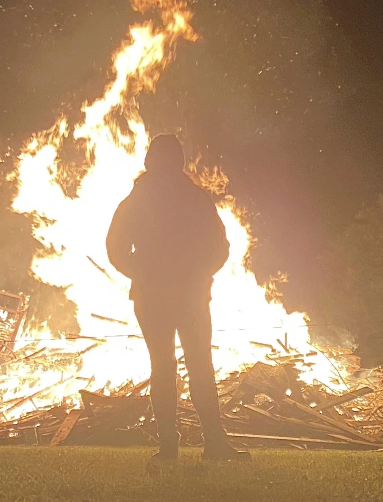 Person in silhouette standing in front of a large bonfire during nighttime.