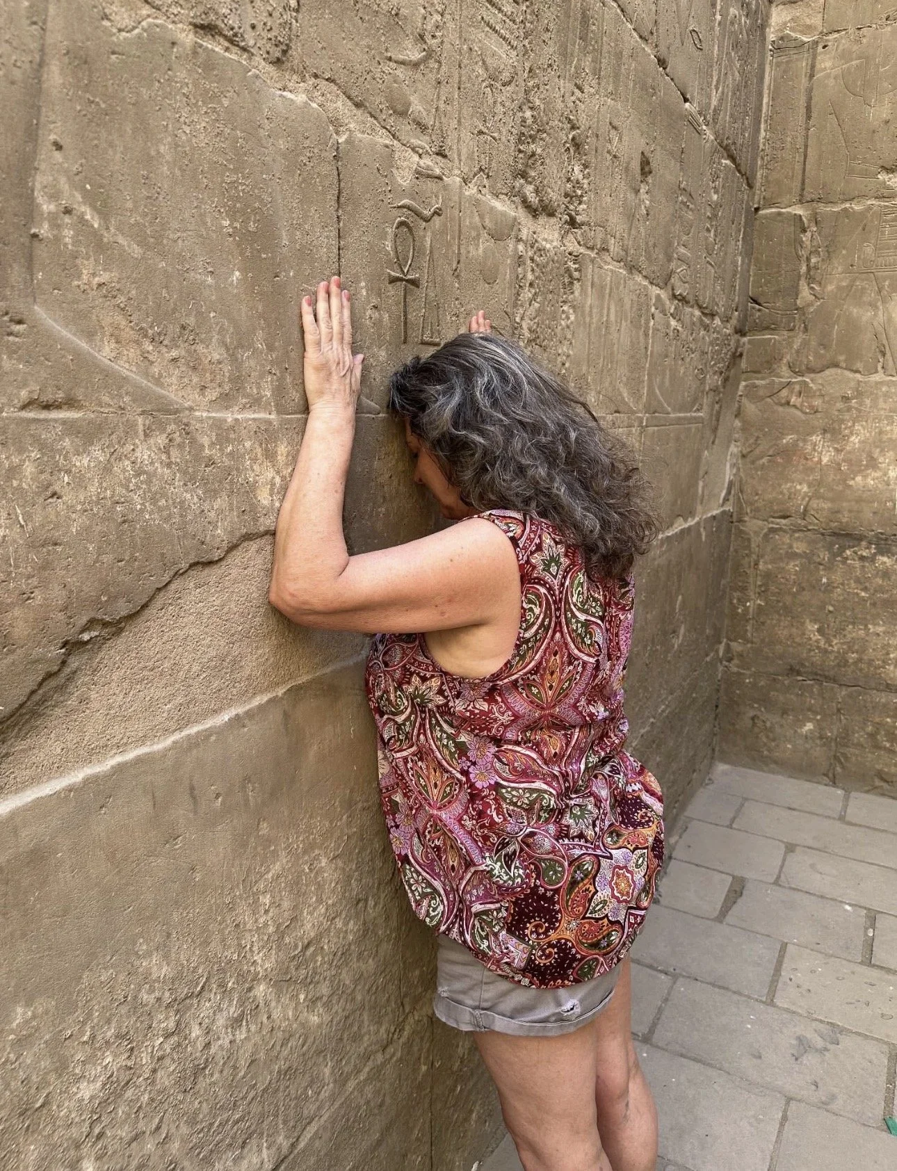 A woman with curly gray hair and tan skin is standing against an ancient stone wall with inscriptions, pressing her ear and hand against it as if listening or reading the carvings.