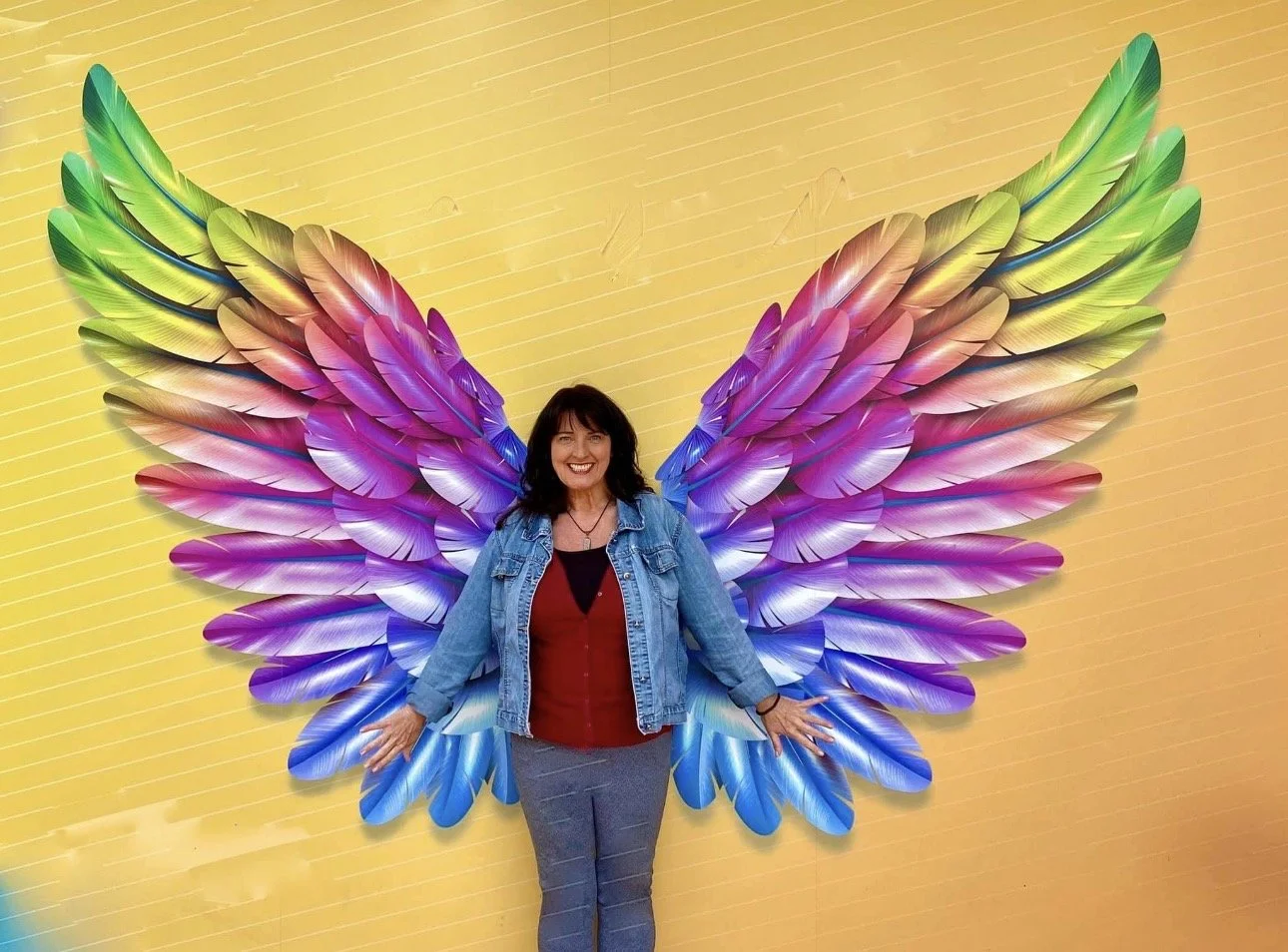 Woman smiling and posing in front of a colorful feathered wing installation on a yellow wall.
