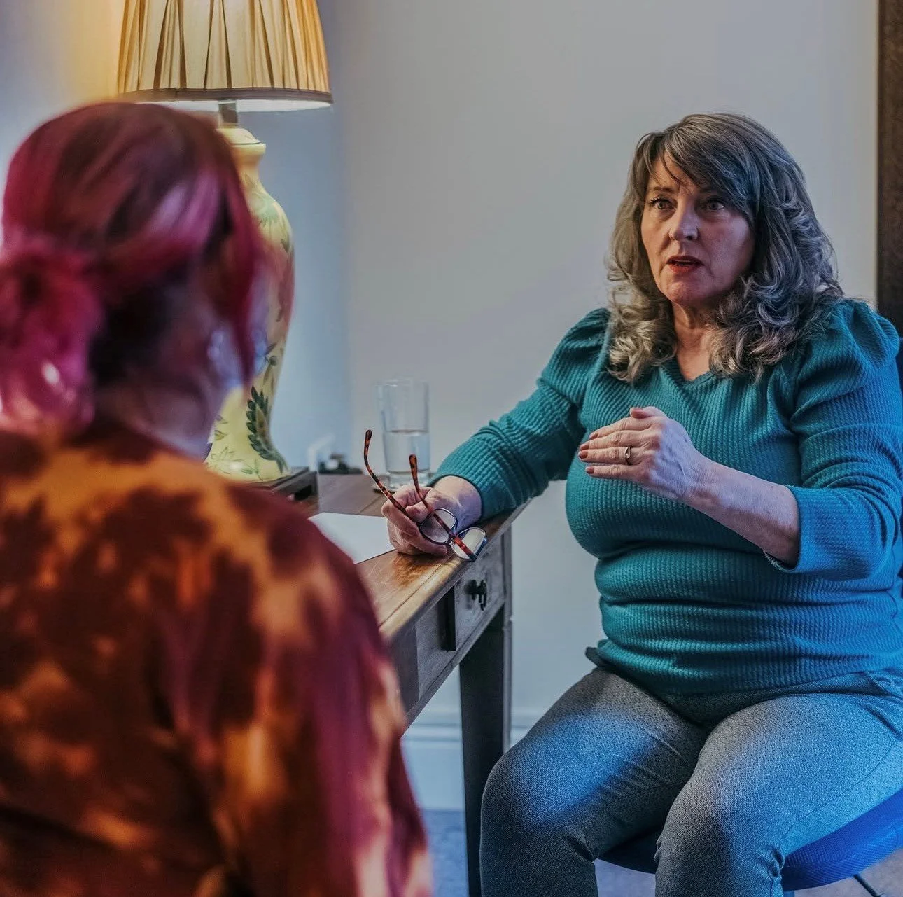 A woman with gray hair and a teal sweater speaking to a person with pink hair in an orange and red top, sitting near a wooden table with a glass of water and eyeglasses in a room with a table lamp.