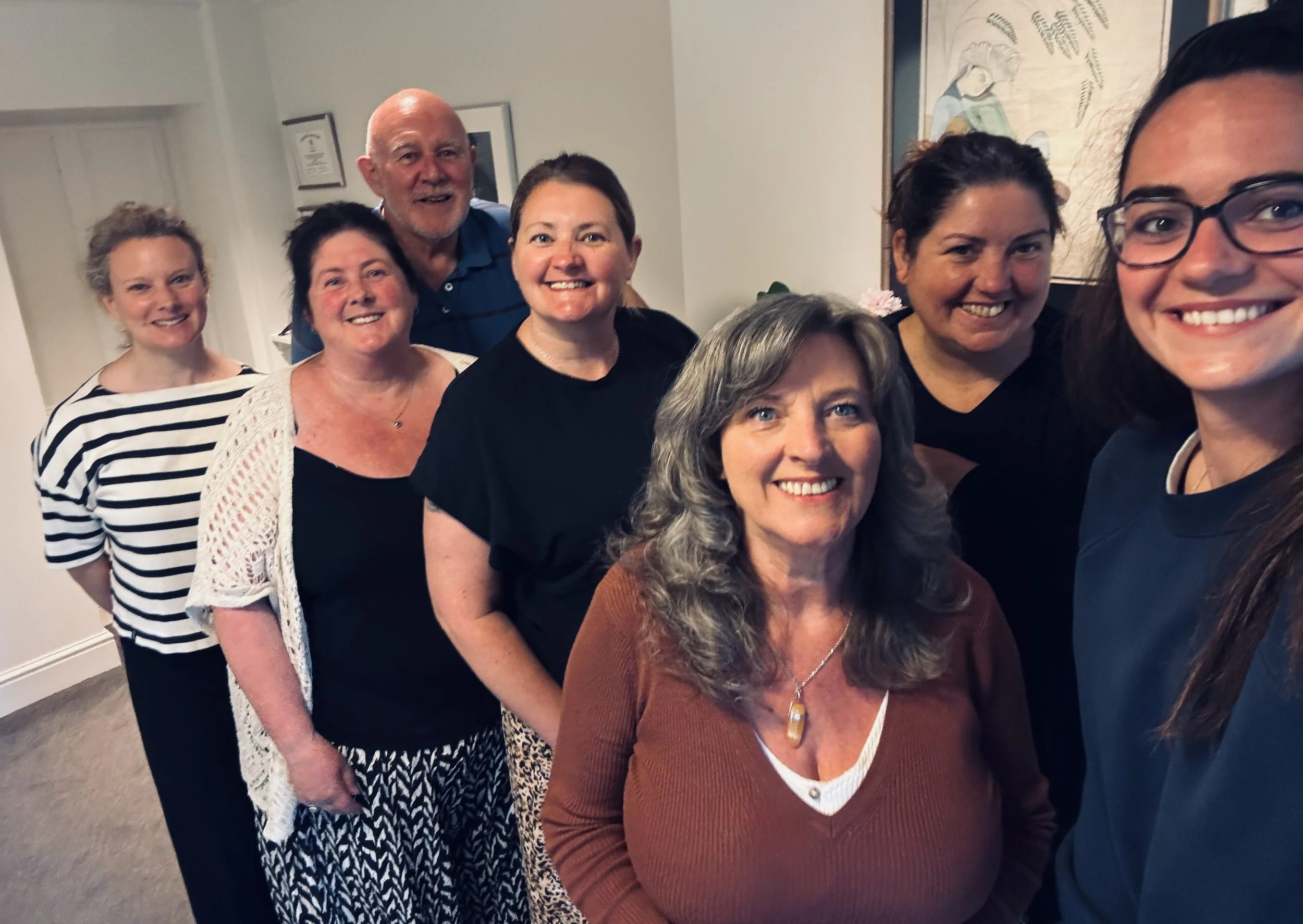 Group of seven smiling people, mostly women, standing close together indoors against a wall with framed pictures and artwork, posing for a photo.