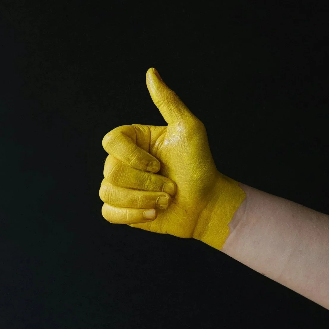 A yellow-painted hand giving a thumbs-up against a dark background, representing creative, hands-on marketing support by Roza Marketing.