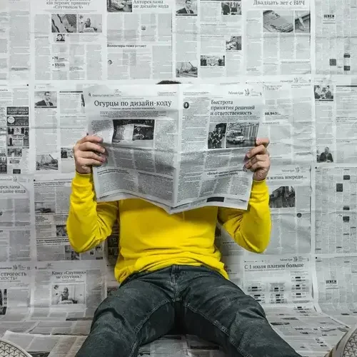 A person in a bright yellow jumper reading a newspaper against a background covered in news print, showcasing traditional marketing and local business advertising strategies at Roza Marketing.