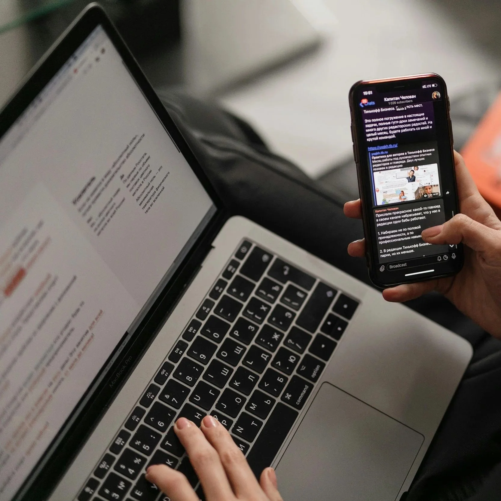 A close up image of a person scrolling their phone and working on their laptop at the same time