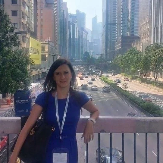 A dark haired woman in a blue dress posing for a picture on a bridge going over a busy road in China