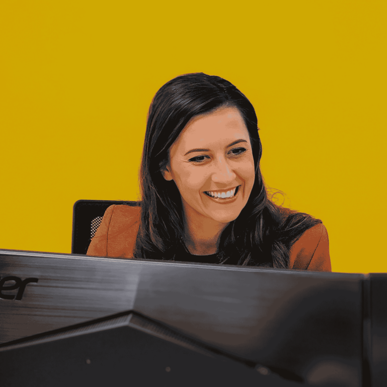 A woman with dark hair smiling at a computer screen against a yellow background.