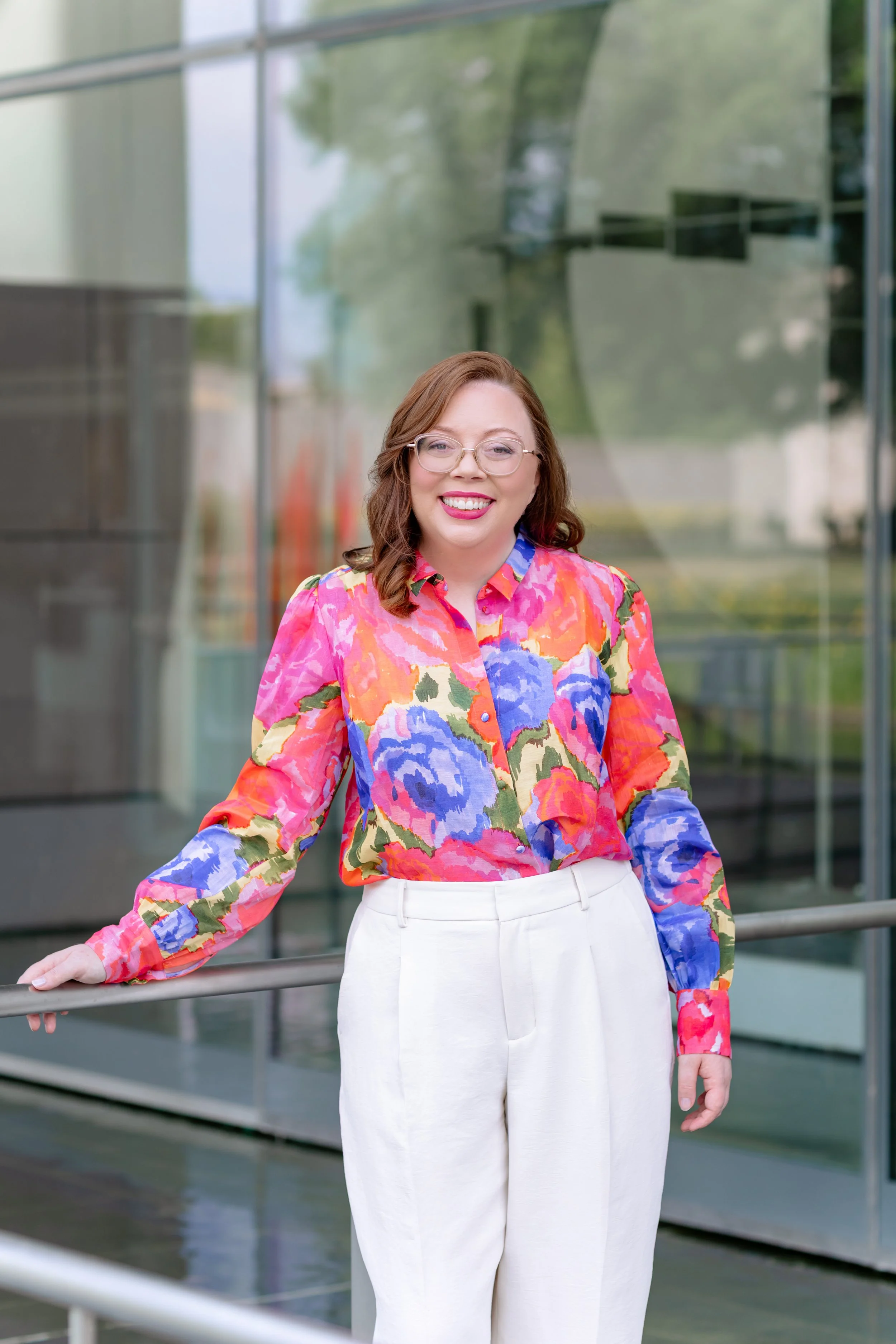 A career coach in Richmond, VA with glasses wearing a colorful floral blouse and white pants standing outside in front of a glass building.