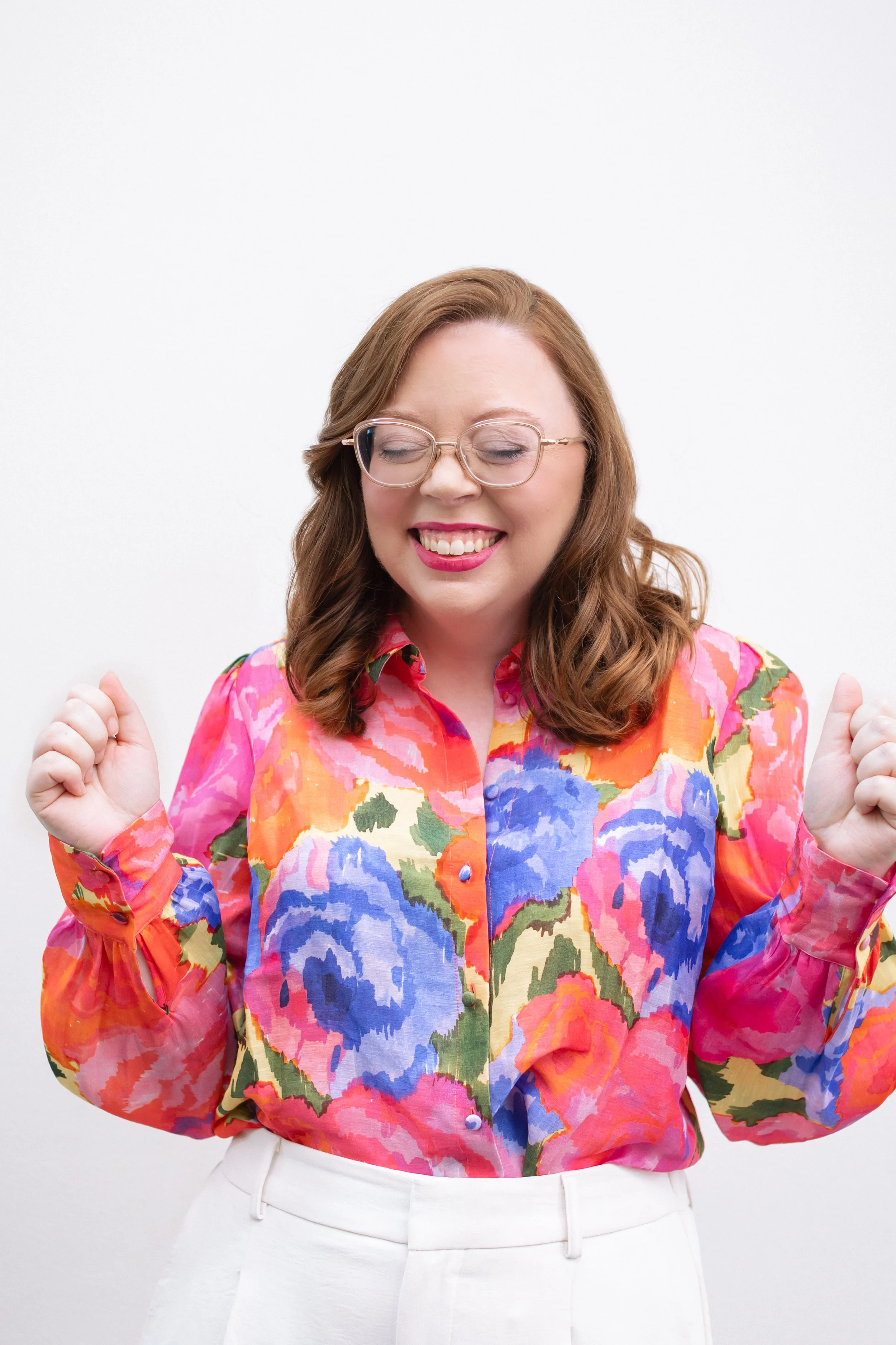 A leadership coach in Richmond, VA smiling with eyes closed, wearing glasses, a multicolored floral blouse, and white pants, against a plain light background.