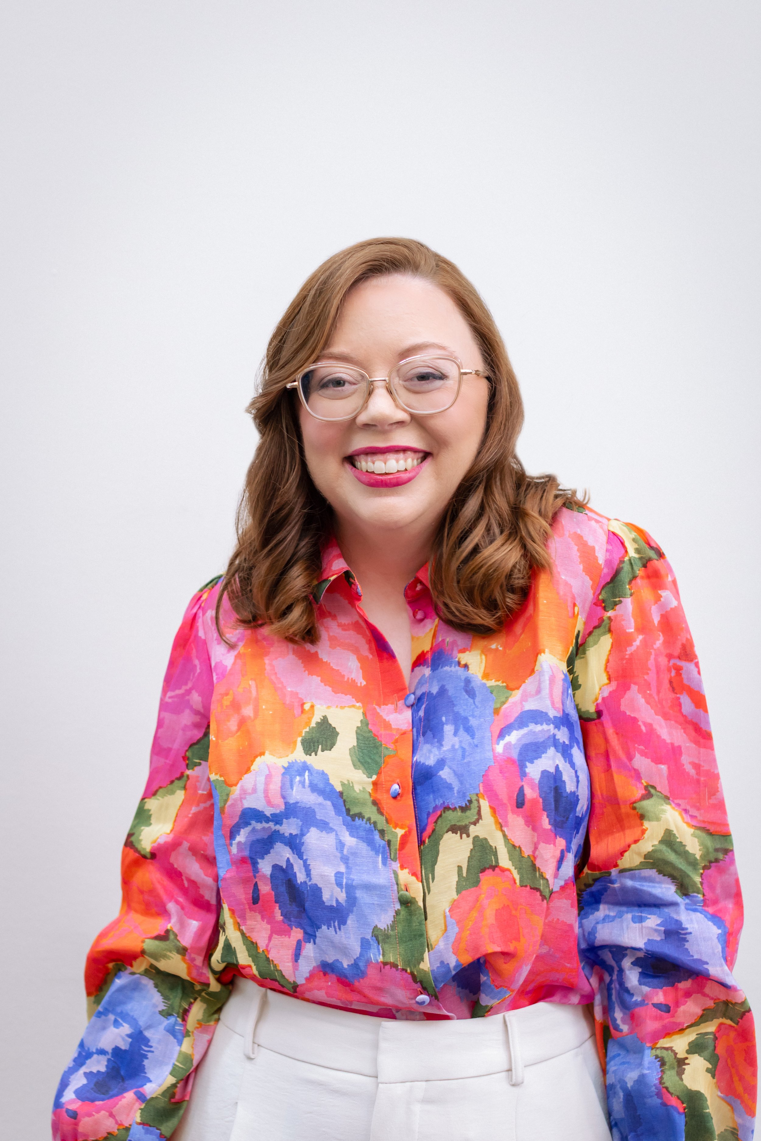 A smiling career coach in Richmond, VA with red hair, glasses, wearing a colorful floral blouse and white pants, standing against a plain white background.