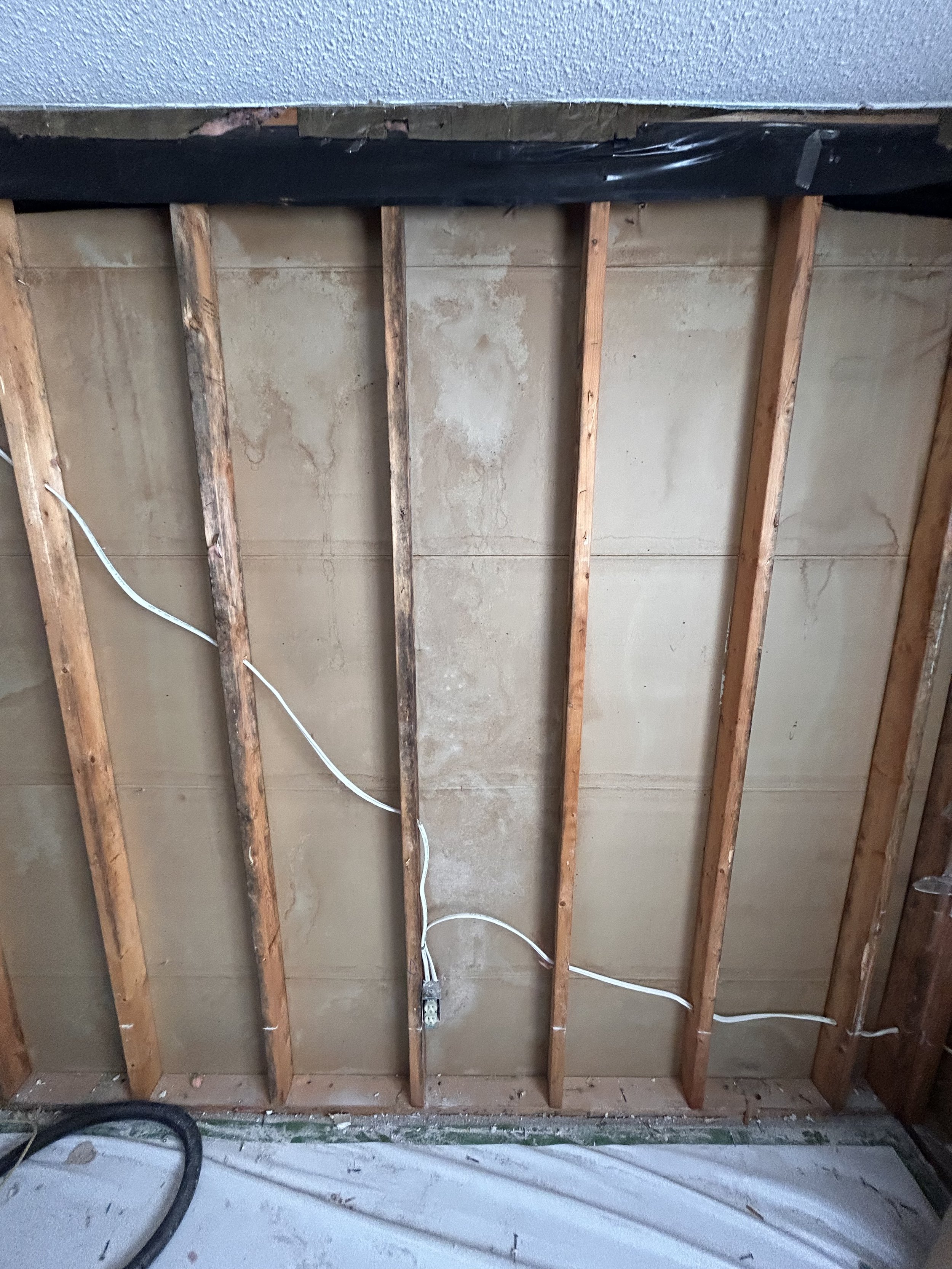View of a wall with exposed wooden studs, electrical wiring, and a concrete wall behind it, indicating construction or renovation work.