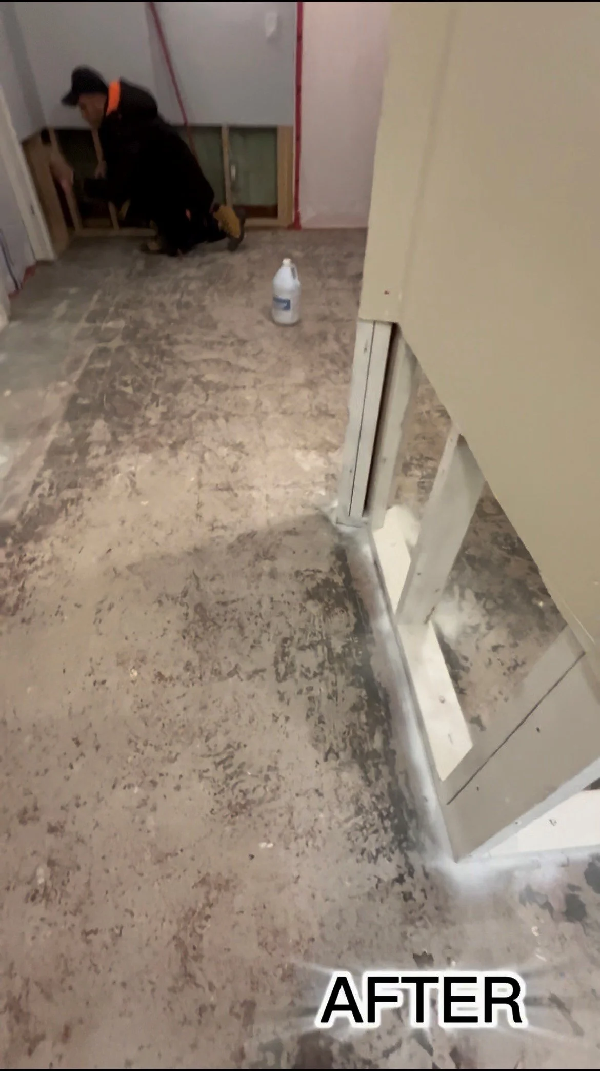 Renovation or construction site showing a partially installed doorway, with a worker kneeling and working on the wall near a window, and a spray bottle placed on the floor.